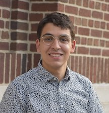 a young man sits smiling straight at the camera against a brick wall