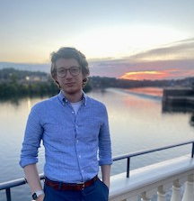 a man wearing glasses stands in front of a lake at sunset