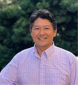 A dark haired man in a pink button-up shirt smiling. 