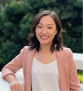 A dark haired woman in a pink blazer smiling in front of trees.