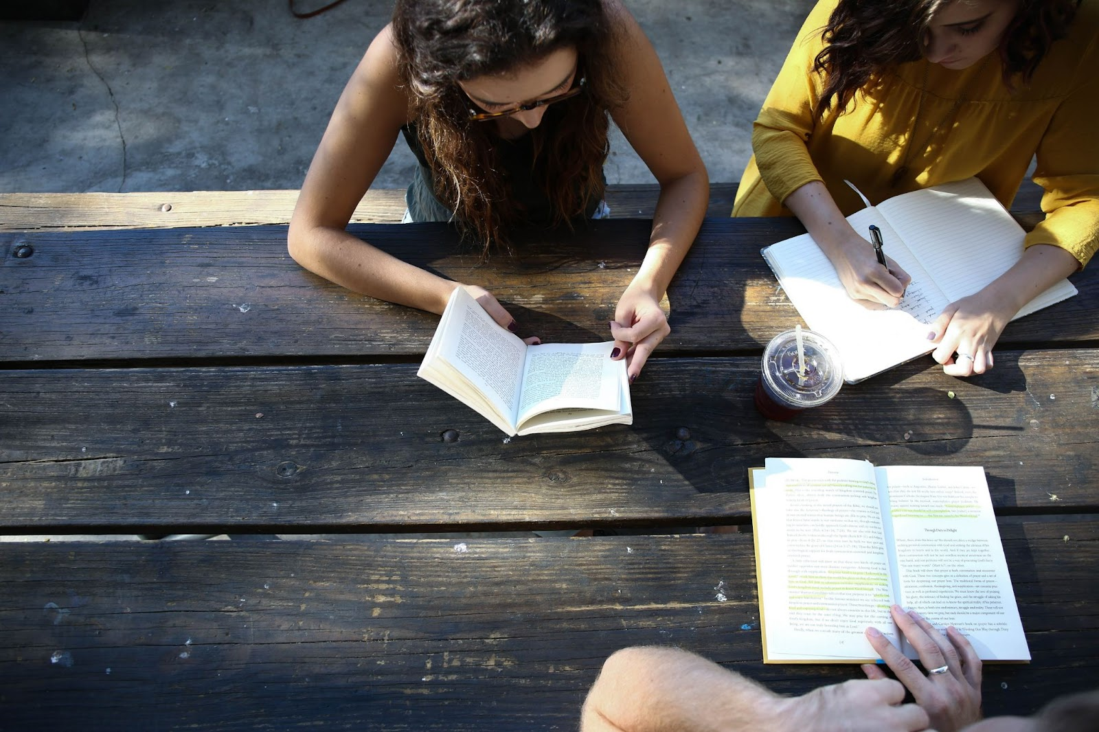 top view down of a wooden table outside, with two people writing things down in a notebook and one person reading