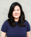 Monica Cho smiling with a stone wall background wearing a dark blue shirt