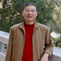 A man with dark hair and glasses stands leaning against a concrete railing wearing a brown jacket and red shirt.