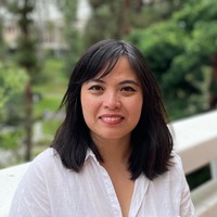 A smiling woman with shoulder-length black hair wearing a white button-up top standing in front of trees.