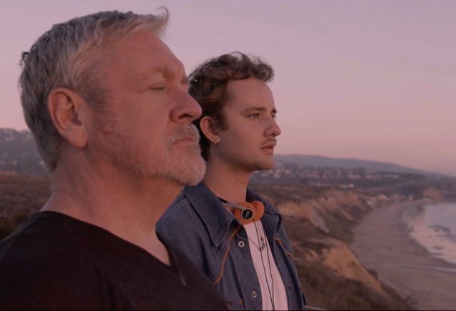 Two men stand on a bluff looking out over the ocean. 