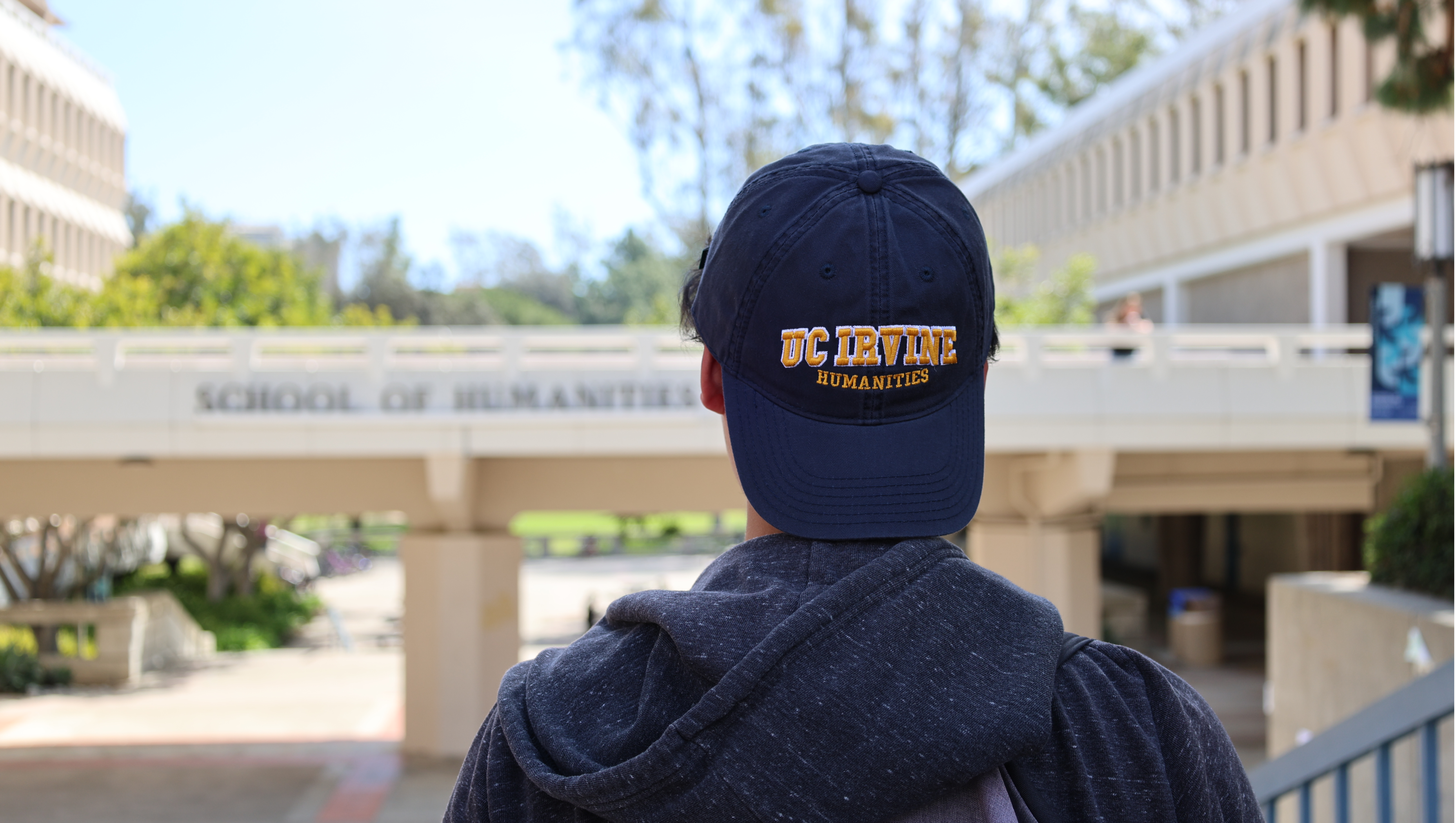 A transfer student with a UC Irvine Humanities baseball cap looking at the School of Humanities bridge.