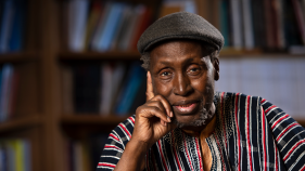 Ngugi rests his head lightly on his left hand; he is in a library