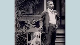 Pale blue book cover with title, An Archive of Skin, An Archive of Kin: Disability and Life-Making During Medical Incarceration. Author Adria L. Imada. The photograph is of a Native Hawaiian man, Naihe Pukai, wearing dark suit, white shirt, and flower in lapel. He stands outside house, looking off to his left. He holds onto wooden chair with right hand. A white dog, at center of photo, sits upright in chair, looking directly at camera.  Kalaupapa, Molokai settlement, ca. 1910.