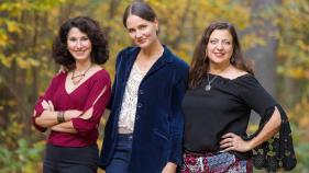 The three women who make up the band Zulal stand next to eachother smiling in front of trees.