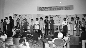 A black and white photo taken at the OCS at a Black History Bee. A group of students stand up on stage in front of an audience.