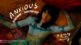 A still from the film ANXIOUS., showing the character Ruby, wearing a color dress, laying in a bed.