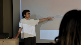 An action shot of UTeach student Brian Nguyen in the classroom talking about his powerpoint slide.