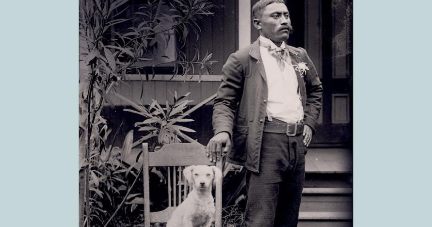 Pale blue book cover with title, An Archive of Skin, An Archive of Kin: Disability and Life-Making During Medical Incarceration. Author Adria L. Imada. The photograph is of a Native Hawaiian man, Naihe Pukai, wearing dark suit, white shirt, and flower in lapel. He stands outside house, looking off to his left. He holds onto wooden chair with right hand. A white dog, at center of photo, sits upright in chair, looking directly at camera.  Kalaupapa, Molokai settlement, ca. 1910.