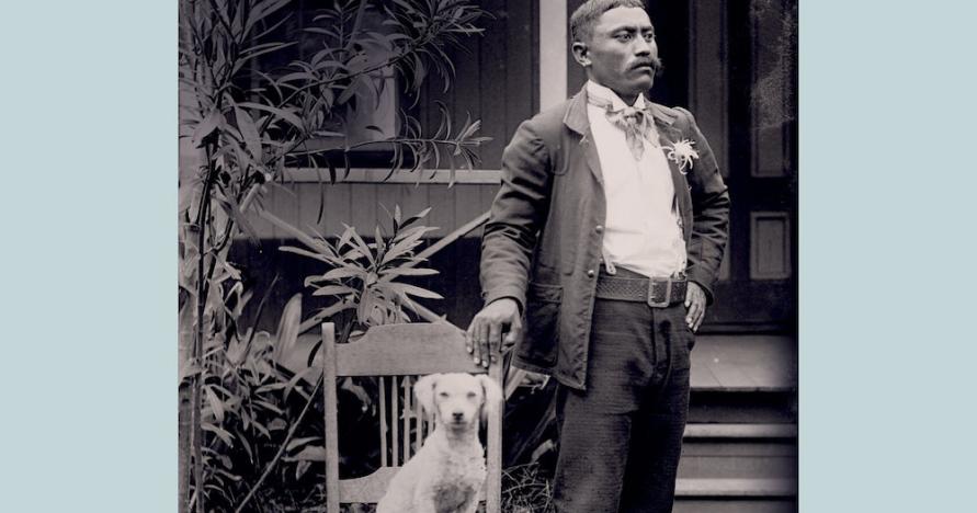 Pale blue book cover with title, An Archive of Skin, An Archive of Kin: Disability and Life-Making During Medical Incarceration. Author Adria L. Imada. The photograph is of a Native Hawaiian man, Naihe Pukai, wearing dark suit, white shirt, and flower in lapel. He stands outside house, looking off to his left. He holds onto wooden chair with right hand. A white dog, at center of photo, sits upright in chair, looking directly at camera. Kalaupapa, Molokai settlement, ca. 1910.