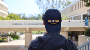 A transfer student with a UC Irvine Humanities baseball cap looking at the School of Humanities bridge.