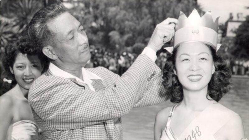 A black and white photo shows a woman being adorned with a crown