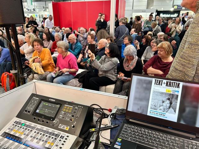 Computer with Text+Kritik book cover with a crowd sitting behind it at the Leipzig Book Fair
