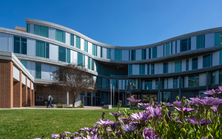 School of Humanities  Building which is waved shaped with blue and white windows. There's a student walking towrds the entrance and there's purple flowers in the foreground.