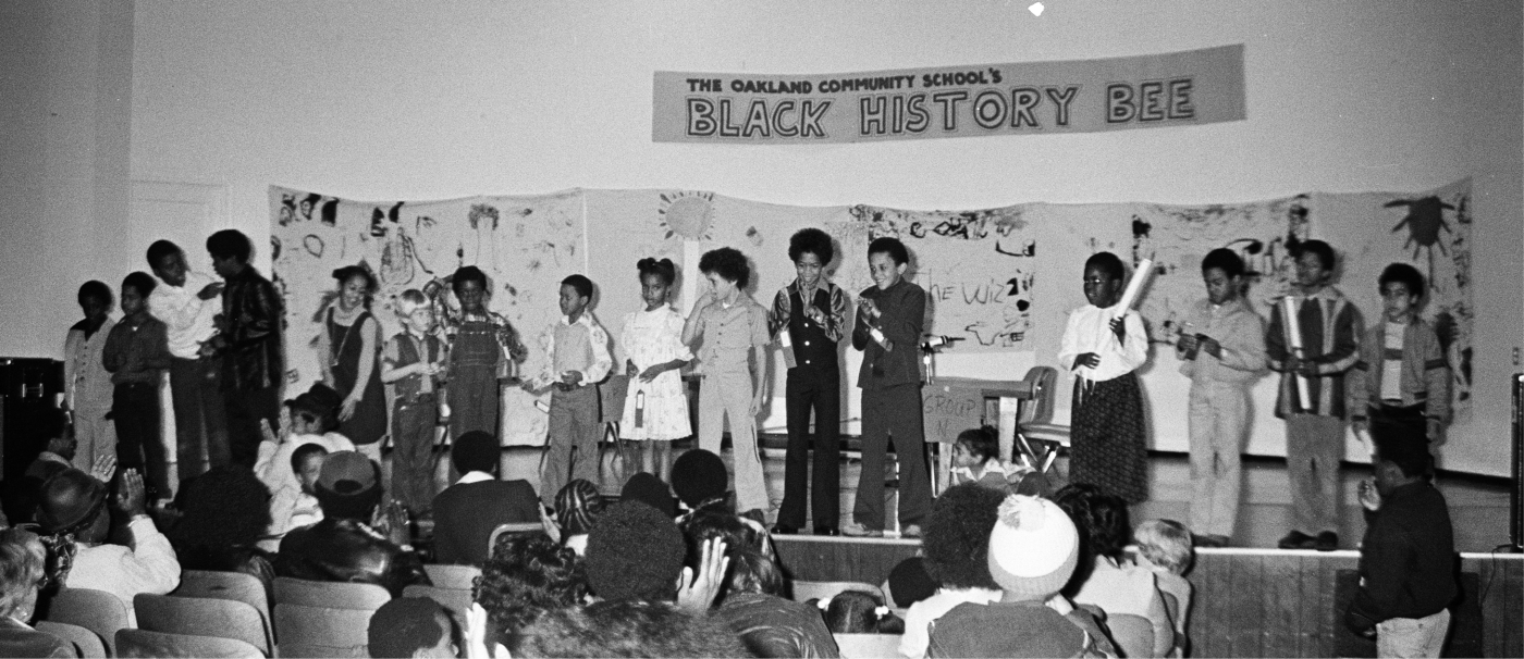 A black and white photo from OCS. Students are standing up on a stage in front of an audience participating the a "Black History Bee".