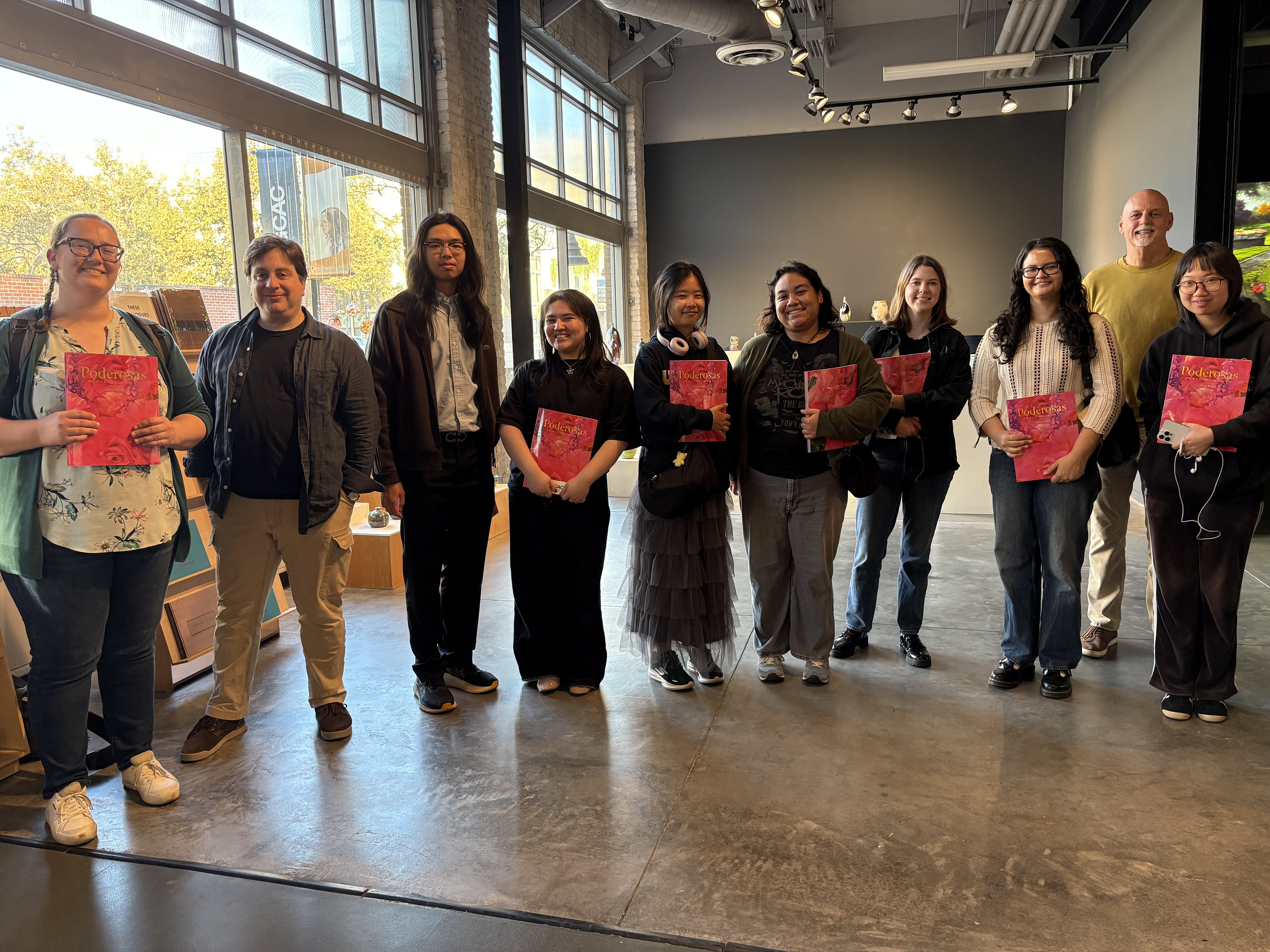 13 people stand together at Grand Central Art Center on Internship Touring Day holding pink books.