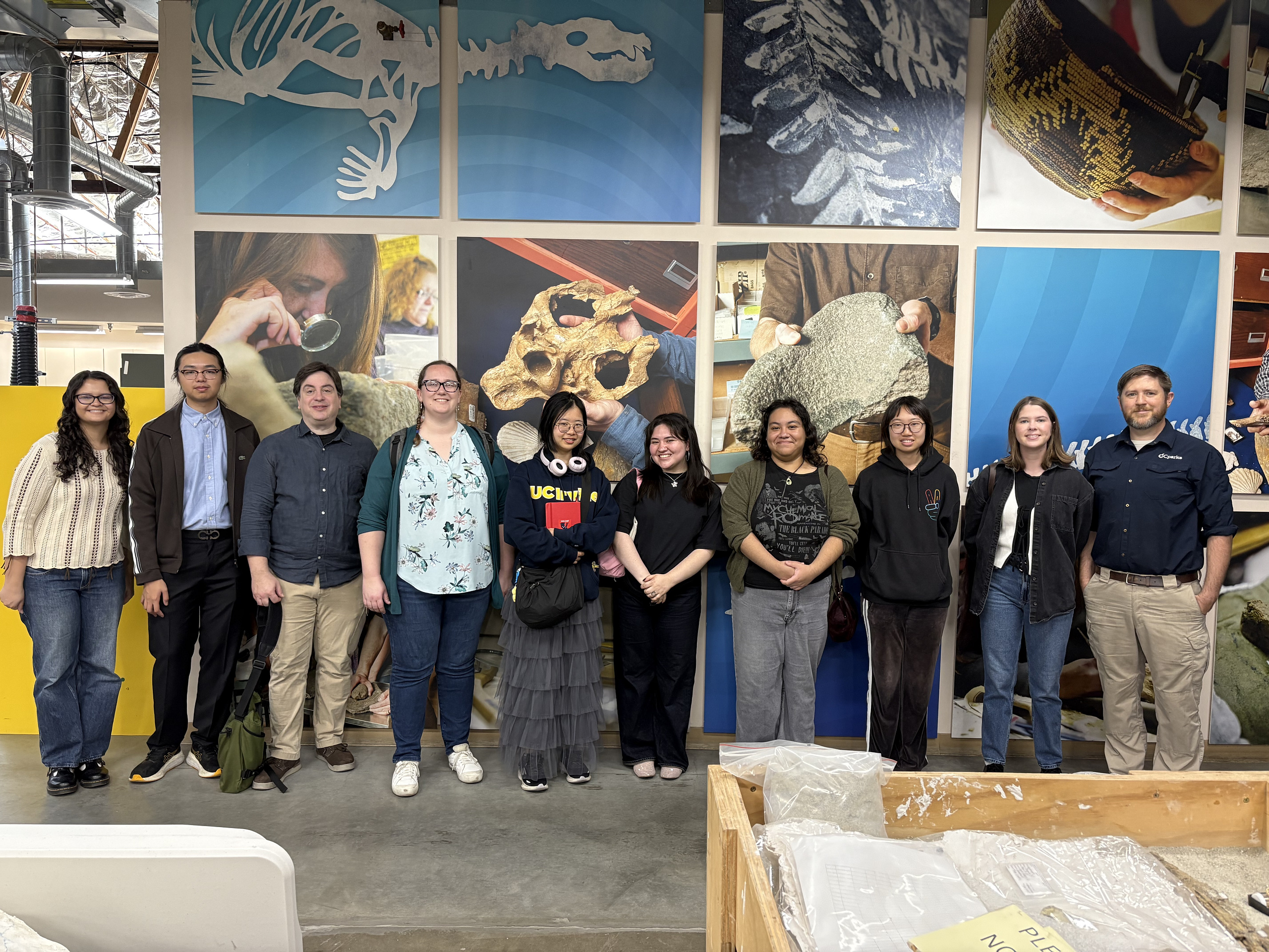 13 people stand together at OC Parks Cooper Lab in front of a paleontology background on Internship Touring Day.
