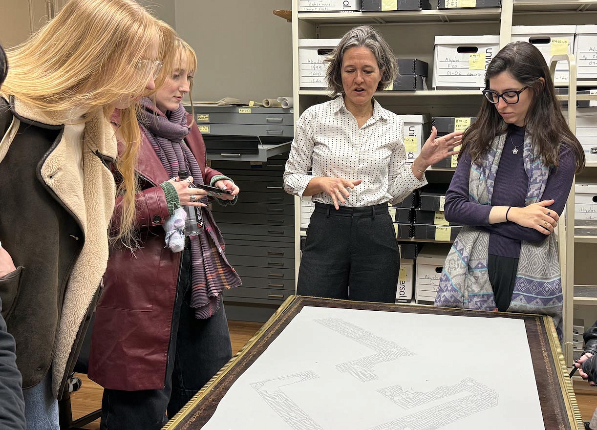 Ellie and her supervisor, Professor Silva, and one other student are standing around a table looking at a large document. 