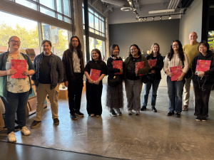13 people stand inside of Grand Central Art Center holding pink books.