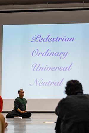 Dancer Gerald Casel seated on floor in front of screen with words Pedestrian. Ordinary. Universal. Neutral.