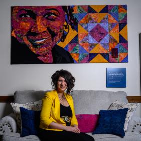 M. Shadee posing in the middle of a couch with paintings above her and a bookcase on either side of the couch