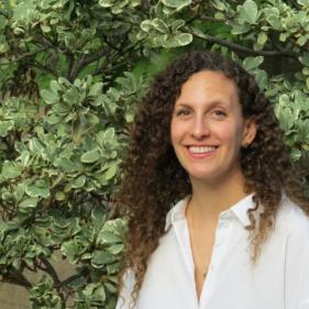 woman with long curly brown hair wearing white shirt in front of green bush