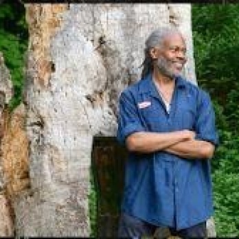Smiling African American man with gray hair wearing a blue shirt and leaning against a tree