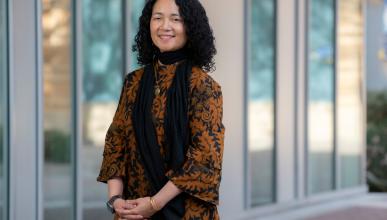 Fatimah Tobing Rony wears a black and orange patterned top and black scarf. She is front of Humanities Gateway at UCI