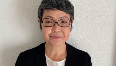 Laura Kang stands against a white stucco wall. She is wearing a black suit coat and white blouse