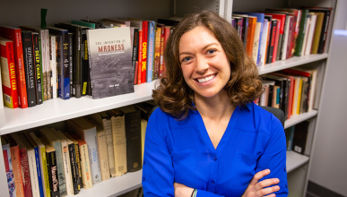 Emily Baum wears royal blue blouse; she stands against a bookshelf with her arms crossed