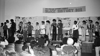 A black and white photo taken at the OCS at a Black History Bee. A group of students stand up on stage in front of an audience.