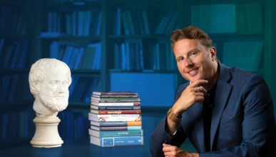 Duncan Pritchard is siting in a library at a table with a pile of books and a philosophers bust. The background has a blue overlay. 