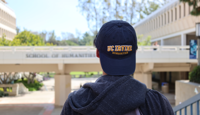 A transfer student with a UC Irvine Humanities baseball cap looking at the School of Humanities bridge.