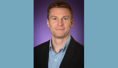Head and shoulders of man with short brown hair, wearing blue shirt and dark jacket